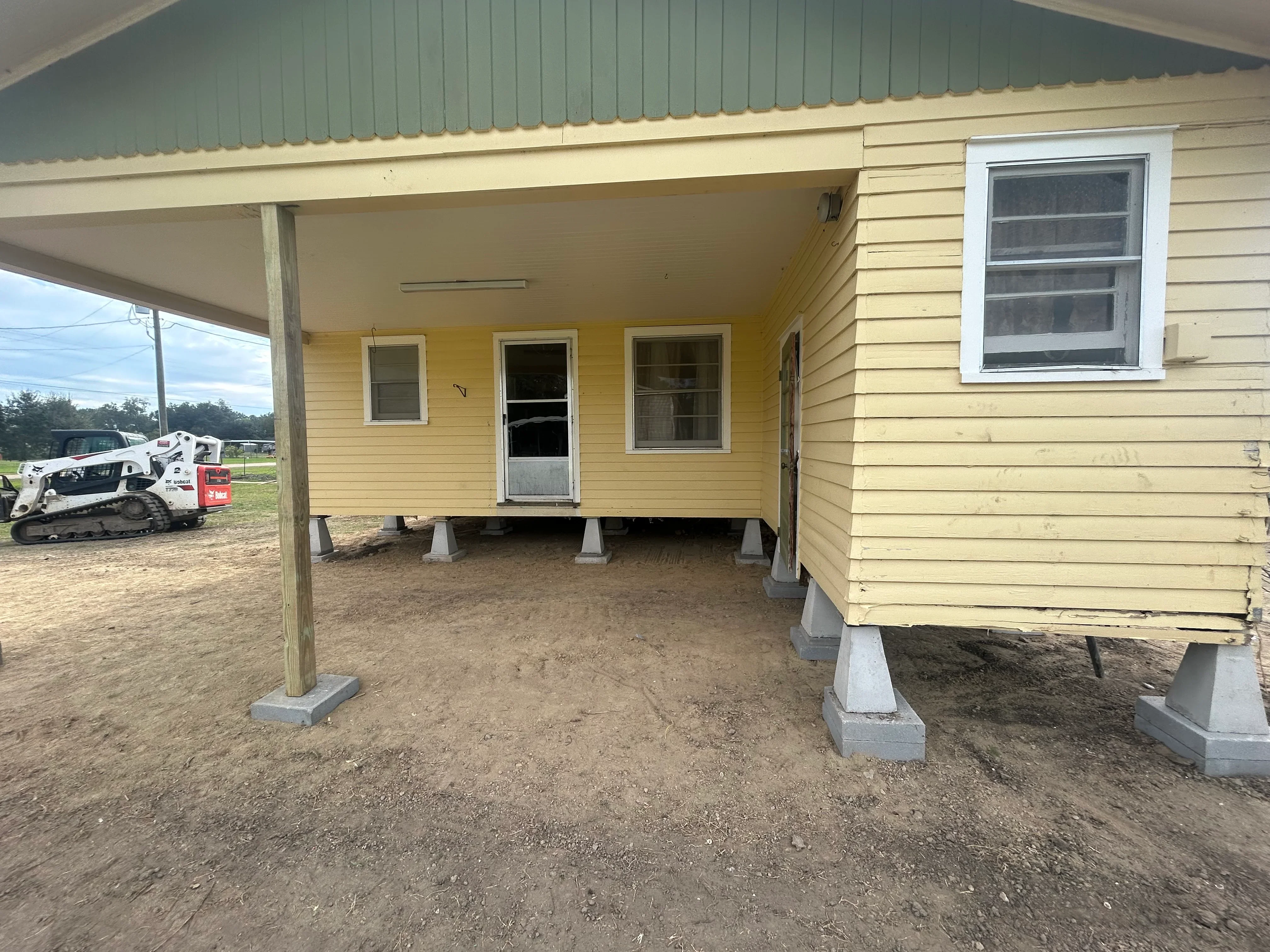 Yellow mobile home elevated on concrete piers and wooden support posts with metal carport